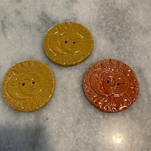 May include: Three circular buttons featuring a raised relief design of a sun and crescent moon face. Two buttons are a mustard yellow color, and one button is a reddish-orange color. Each button has two small holes in the center for attachment.