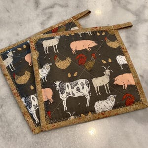 May include: Two brown and tan quilted pot holders with a farmhouse theme. The pot holders feature a black background with white and pink farm animals, including cows, pigs, sheep, chickens, and goats.
