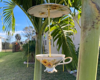 May include: A decorative bird feeder made from a teacup and saucer, suspended from a gold-colored metal hanger. The teacup and saucer are white with a yellow sunflower design. The feeder is hanging from a palm tree in a sunny outdoor setting.