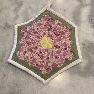 May include: A quilted, floral-patterned hot pad in a starburst design. The center features a yellow, quilted accent. The main pattern is a light cream color with purple floral designs. The edges are bordered with green and white fabric.