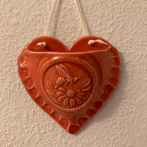 May include: A heart-shaped, coral-colored ceramic wall pocket with a floral and bee relief design. The pocket has a scalloped edge and a white ribbon for hanging. The design features a bee and a daisy.