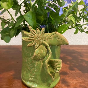 May include: A cylindrical green ceramic vase featuring a raised, sculpted leaf and vine design on its exterior. The vase is filled with a bouquet of vibrant blue flowers and green foliage, showcasing a textured surface with earthy brown accents.