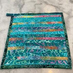 May include: A teal blue quilted hot pad with a colorful batik pattern. The hot pad is made of fabric strips sewn together in a horizontal pattern.