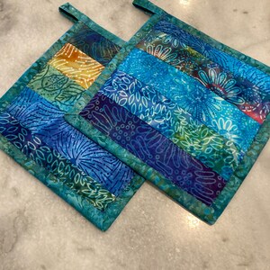 May include: Two square quilted potholders featuring a patchwork design of batik fabrics in shades of blue, teal, purple, and yellow. Each potholder has a fabric loop at the top corner for hanging and is finished with a teal fabric binding border.