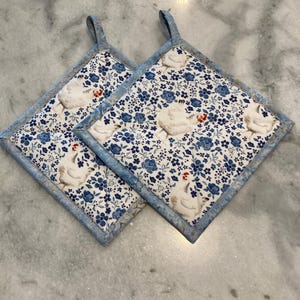May include: Two square pot holders with a blue floral and chicken pattern. The pot holders have a light blue border and a loop for hanging. The fabric features white chickens and small blue flowers.