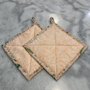 May include: Two square quilted pot holders. The pot holders have a peach-colored background with a white leaf pattern. The edges are trimmed with a floral print and a small loop for hanging.