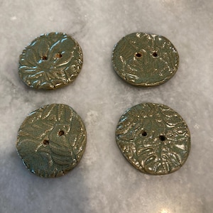 May include: Four round, sage green ceramic buttons with a glossy finish and a textured leaf pattern embossed on the surface. Each button features two central holes for attachment, showcasing a slightly irregular, hand-formed circular shape.