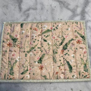 May include: A rectangular, quilted placemat with a floral pattern in shades of pink, white, and green on a peach background. The placemat has a light green border.