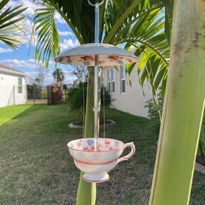 May include: A decorative bird feeder made from a vintage teacup and saucer. The white teacup has a floral pattern with red and blue accents. The saucer and teacup are suspended from a metal rod, creating a charming garden accent.