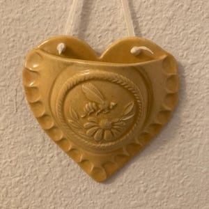 May include: A heart-shaped, yellow ceramic wall pocket with a bee and flower design. The pocket has a scalloped edge and a rope-like border around the central design. A white string is attached for hanging.