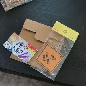 Pu&ograve; includere: Un sacchetto di plastica trasparente pieno di vari adesivi e una busta di carta marrone. Gli adesivi includono un'emoticon gialla sorridente, un fiore viola, una scala con la scritta "Seminterrato" e un adesivo giallo con il testo "Good Vibes Only".