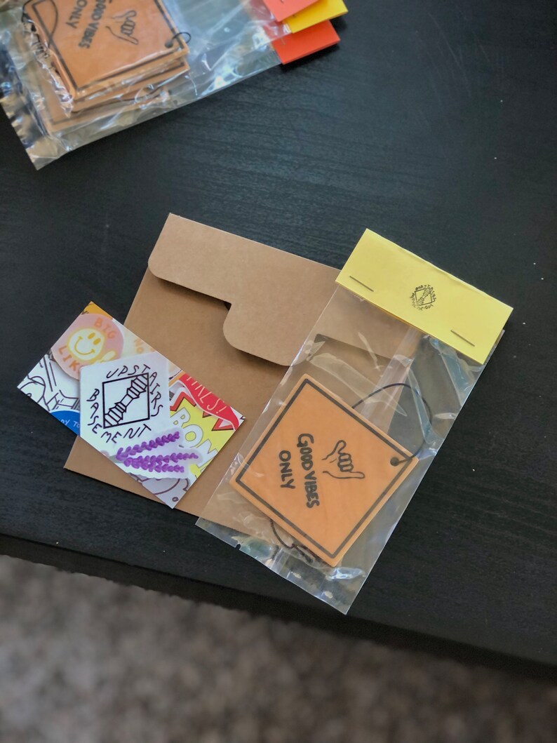 Pu&ograve; includere: Un sacchetto di plastica trasparente pieno di vari adesivi e una busta di carta marrone. Gli adesivi includono un'emoticon gialla sorridente, un fiore viola, una scala con la scritta "Seminterrato" e un adesivo giallo con il testo "Good Vibes Only".