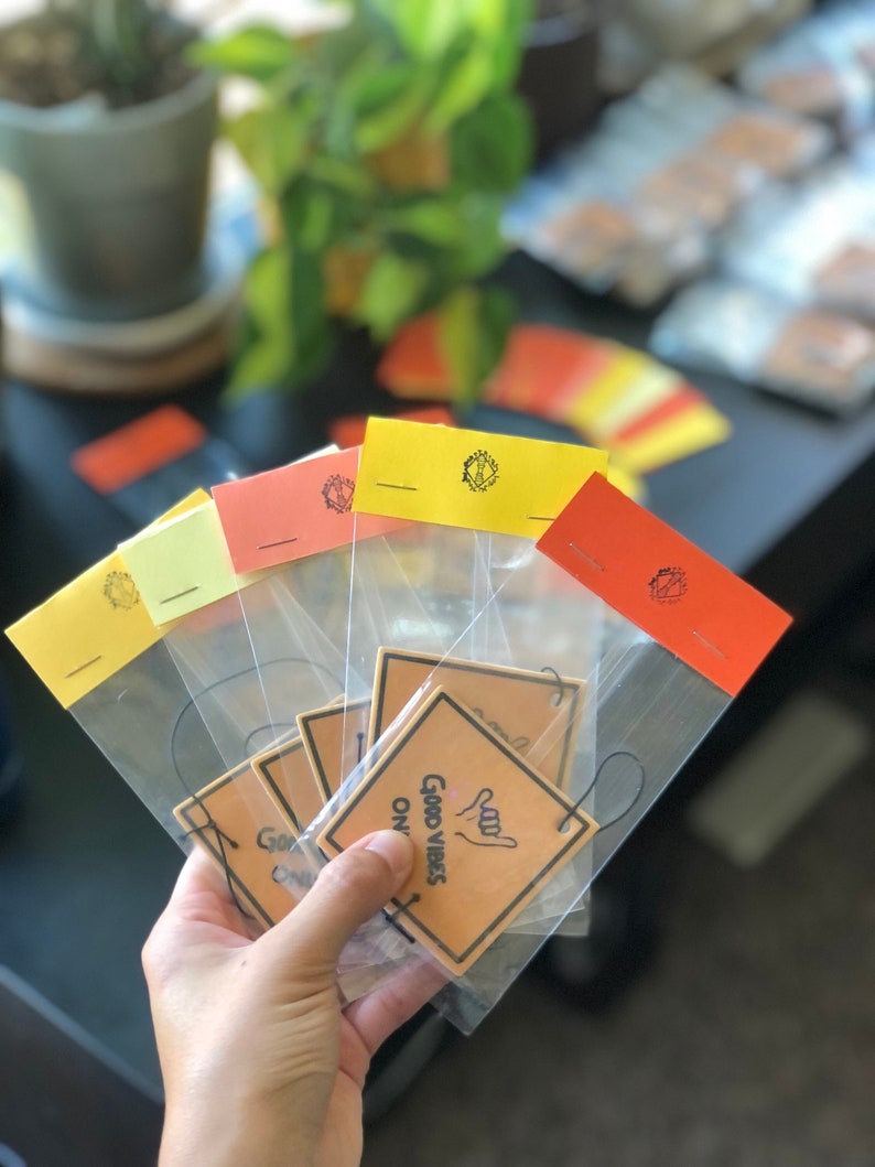 Pu&ograve; includere: Un set di cinque profumatori per auto in sacchetti di plastica trasparente. I profumatori sono marroni con testo nero che recita "Good Vibes Only" e un'immagine disegnata a mano di una mano che fa il segno shaka. I sacchetti sono sigillati con un adesivo giallo, arancione o rosso.