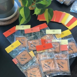 Pu&ograve; includere: Primo piano di una pila di profumatori per auto in carta marrone con il testo "Good Vibes Only" e un'icona disegnata a mano di una mano che fa il segno shaka. I profumatori sono confezionati in sacchetti di plastica trasparente con etichette gialle, rosse e bianche.