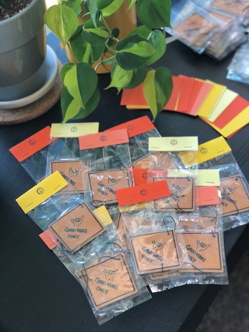 Pu&ograve; includere: Primo piano di una pila di profumatori per auto in carta marrone con il testo "Good Vibes Only" e un'icona disegnata a mano di una mano che fa il segno shaka. I profumatori sono confezionati in sacchetti di plastica trasparente con etichette gialle, rosse e bianche.