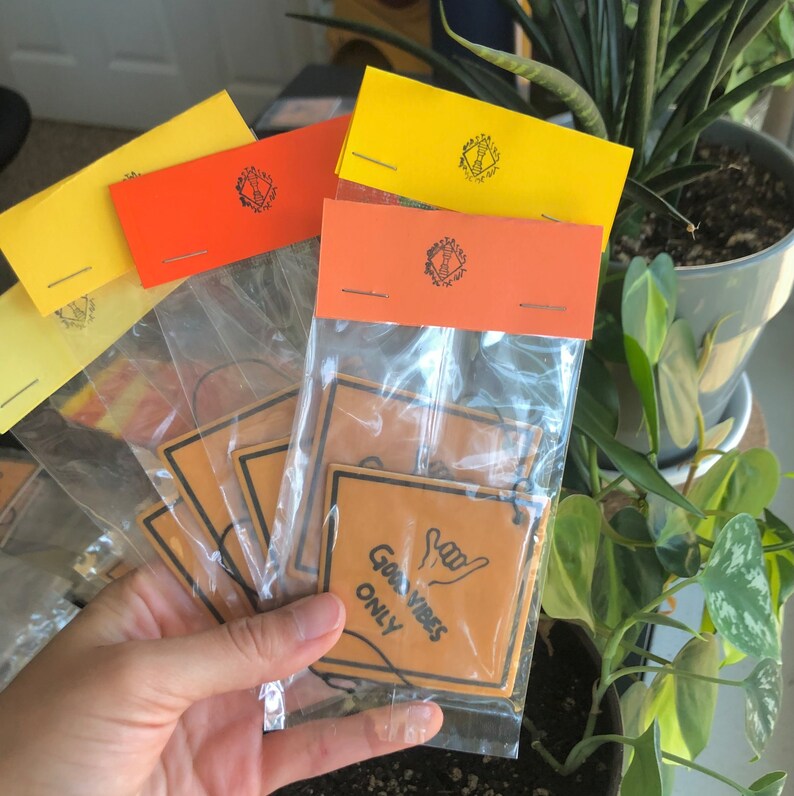 Pu&ograve; includere: Un set di cinque profumatori per auto in giallo, arancione e marrone. I profumatori presentano un gesto della mano e il testo "Good Vibes Only".