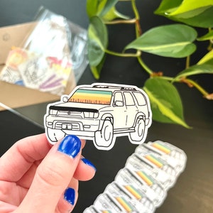 May include: A white car sticker with a black outline. The car has a rainbow colored mountain scene on the back window. The sticker is being held by a hand with blue nail polish.