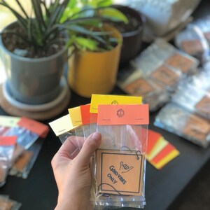 Pu&ograve; includere: Un set di quattro profumatori per auto in carta marrone con un laccio per appenderli. I profumatori sono confezionati in sacchetti di plastica trasparente. La parte anteriore del profumatore dice "Good Vibes Only" in testo nero con un'immagine disegnata a mano di una mano che fa il segno shaka.