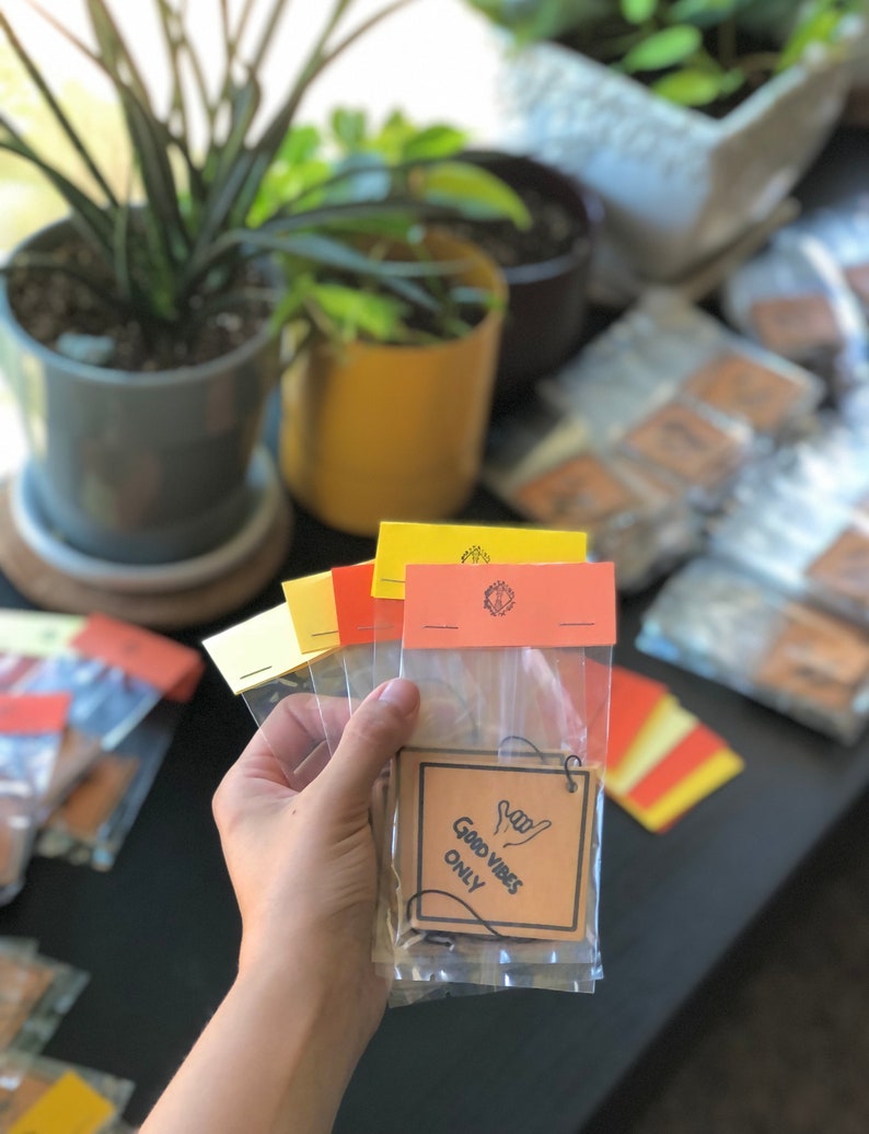 Pu&ograve; includere: Un set di quattro profumatori per auto in carta marrone con un laccio per appenderli. I profumatori sono confezionati in sacchetti di plastica trasparente. La parte anteriore del profumatore dice "Good Vibes Only" in testo nero con un'immagine disegnata a mano di una mano che fa il segno shaka.