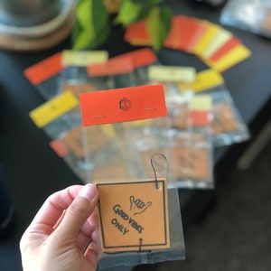 Pu&ograve; includere: Un profumatore per auto di colore beige con uno spago nero appeso in cima. Il profumatore ha un disegno a mano nero di una mano che fa il segno 'hang loose' e il testo 'Good Vibes Only'.