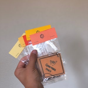 Pu&ograve; includere: Un profumatore per auto in cartone marrone con uno spago nero appeso in cima. Il profumatore ha la scritta "Good Vibes Only" e un'immagine disegnata a mano di una mano che fa il segno "hang loose".