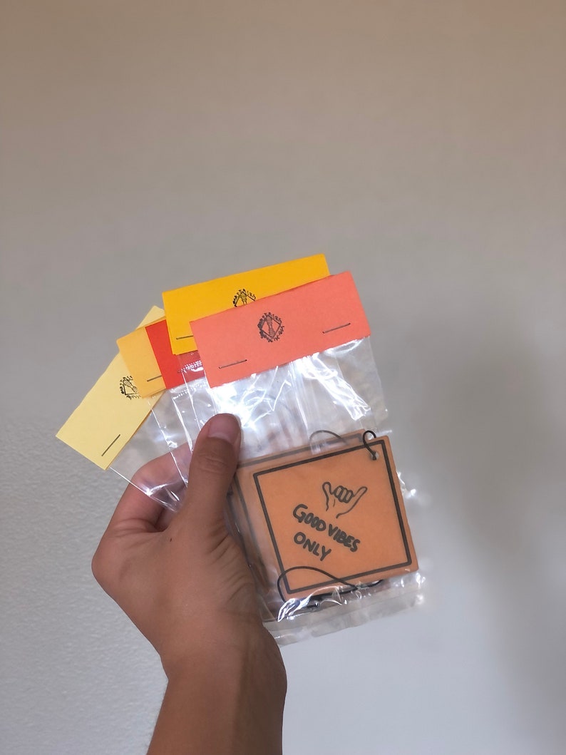 Pu&ograve; includere: Un profumatore per auto in cartone marrone con uno spago nero appeso in cima. Il profumatore ha la scritta "Good Vibes Only" e un'immagine disegnata a mano di una mano che fa il segno "hang loose".