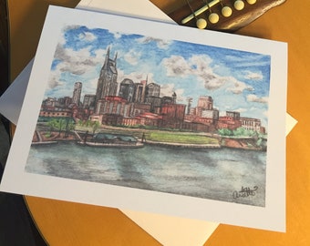 May include: A watercolor print of the Nashville, Tennessee skyline, featuring the city's buildings and the Cumberland River. The artwork is on a white card with an envelope, resting on a wooden acoustic guitar.