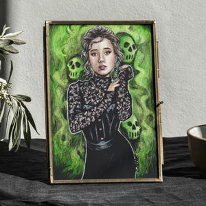 Puede incluir: Una pintura al agua enmarcada de una mujer con un vestido negro de encaje sobre un fondo verde. La mujer tiene el pelo oscuro y está rodeada de calaveras verdes.