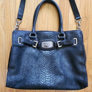 May include: A black handbag with a textured snakeskin pattern, featuring two top handles and a detachable shoulder strap with silver-tone chain links. The front includes a decorative horizontal strap with silver buckles and a central metal logo plate.