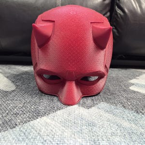 May include: A red mask with a textured surface, featuring two prominent horns and eye openings. The mask has a distinct design, reminiscent of a superhero costume. The mask is likely a cosplay or costume accessory.