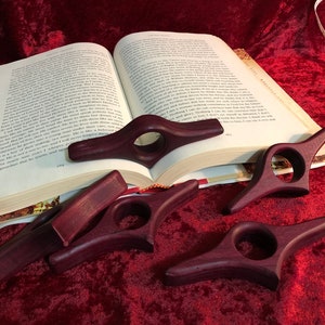 Puede incluir: Varios sujetalibros de madera de color p&uacute;rpura oscuro hechos a mano se exhiben sobre una superficie de terciopelo rojo junto a un libro abierto. Cada soporte presenta un orificio para el dedo y un dise&ntilde;o curvo para mantener las p&aacute;ginas abiertas. El libro est&aacute; abierto en la p&aacute;gina 164.