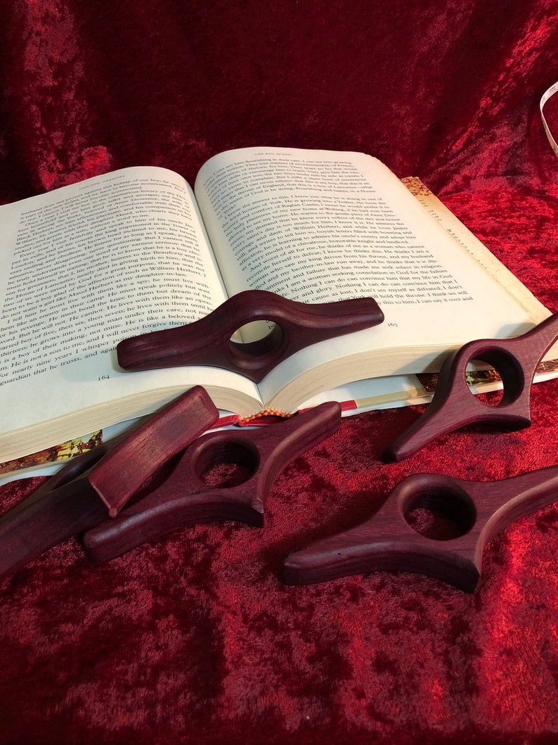 Puede incluir: Varios sujetalibros de madera de color p&uacute;rpura oscuro hechos a mano se exhiben sobre una superficie de terciopelo rojo junto a un libro abierto. Cada soporte presenta un orificio para el dedo y un dise&ntilde;o curvo para mantener las p&aacute;ginas abiertas. El libro est&aacute; abierto en la p&aacute;gina 164.