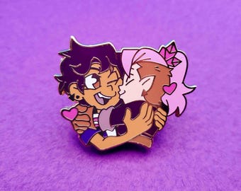May include: A gold-coloured enamel pin featuring two cartoon characters, a purple-haired person with brown skin and a pink-haired person with brown skin, hugging and kissing. The pin has pink hearts around the characters.