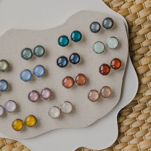 Op de afbeelding: Een verzameling van 10 paar regenboogkleurige ronde oorbelletjes, gepresenteerd op een wit en beige oppervlak. De oorbelletjes zijn in rijen gerangschikt en tonen een verscheidenheid aan kleuren, waaronder blauw, groen, roze, rood en geel.