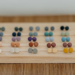 May include: A collection of 20 pairs of small, round, colorful clay earrings displayed on a wooden board. The earrings are arranged in rows of two, with each pair featuring a different color or pattern.