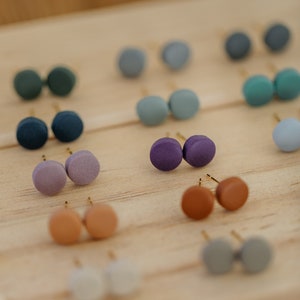 May include: A display of small, round, colorful earrings on a wooden surface. The earrings are arranged in pairs and come in a variety of colors, including purple, blue, green, brown, and gray.