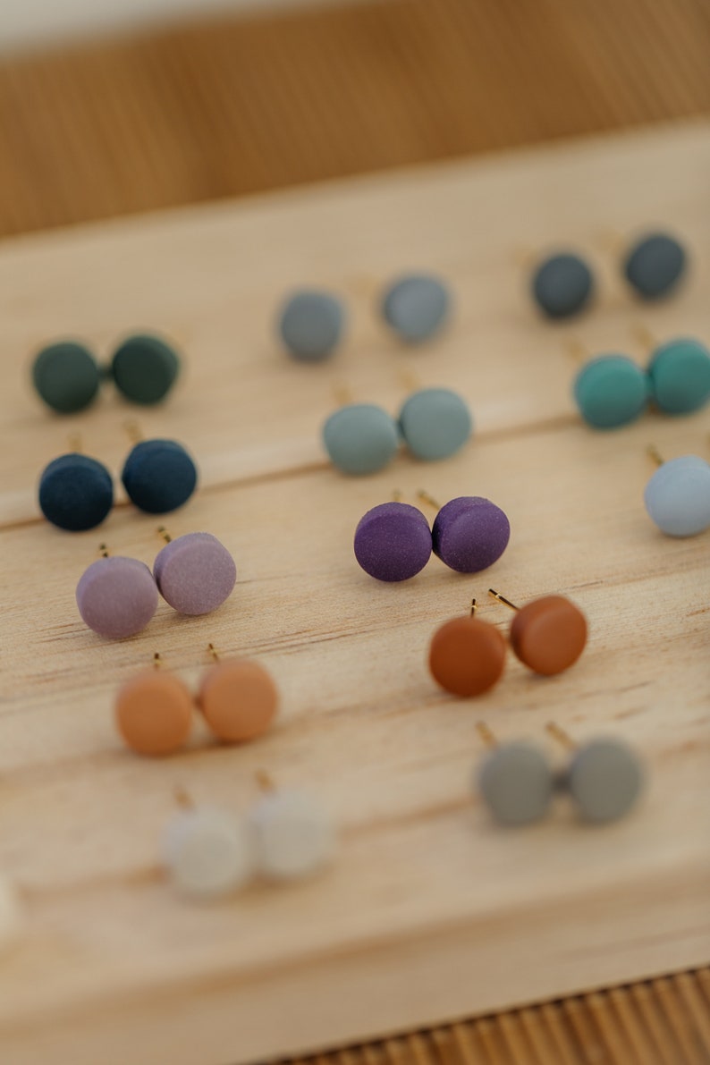 May include: A display of small, round, colorful earrings on a wooden surface. The earrings are arranged in pairs and come in a variety of colors, including purple, blue, green, brown, and gray.