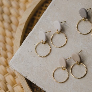 May include: Three pairs of gold hoop earrings with small beige and gray round ceramic accents. The earrings are displayed on a light gray textured surface.