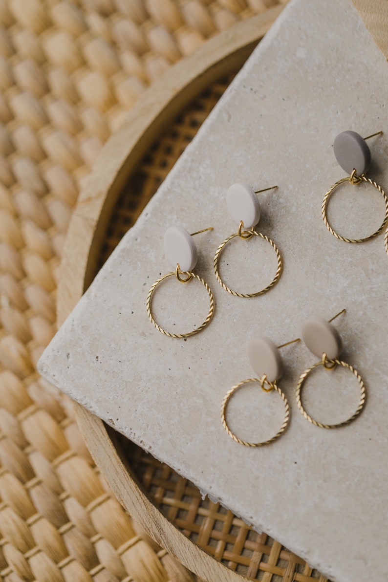 May include: Three pairs of gold hoop earrings with small beige and gray round ceramic accents. The earrings are displayed on a light gray textured surface.