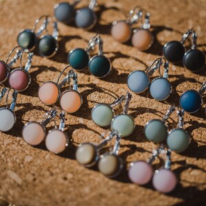 May include: Assortment of colorful round glass earrings with silver hooks. The earrings are displayed on a cork surface.