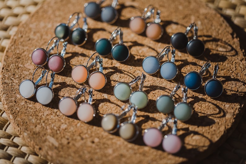 May include: Assortment of colorful round glass earrings with silver hooks. The earrings are displayed on a cork surface.