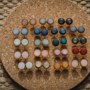 Pu&ograve; includere: Un pannello di sughero con una variet&agrave; di orecchini rotondi e colorati esposti su di esso. Gli orecchini sono disposti in file e sono di diversi colori, tra cui blu, verde, rosso, rosa e giallo.