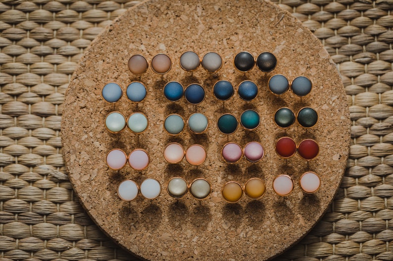 Pu&ograve; includere: Un pannello di sughero con una variet&agrave; di orecchini rotondi e colorati esposti su di esso. Gli orecchini sono disposti in file e sono di diversi colori, tra cui blu, verde, rosso, rosa e giallo.