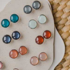 Op de afbeelding: Assortiment van kleurrijke ronde oorbel studs op een witte achtergrond. De oorbel studs zijn gemaakt van metaal en hebben verschillende kleuren, waaronder blauw, groen, rood, roze en wit.