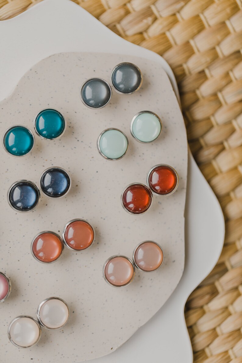 Op de afbeelding: Assortiment van kleurrijke ronde oorbel studs op een witte achtergrond. De oorbel studs zijn gemaakt van metaal en hebben verschillende kleuren, waaronder blauw, groen, rood, roze en wit.