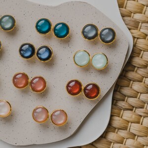 May include: Assortment of colorful round gemstone earrings displayed on a white ceramic plate. The earrings are gold-toned and feature various colors, including blue, green, red, pink, and white.