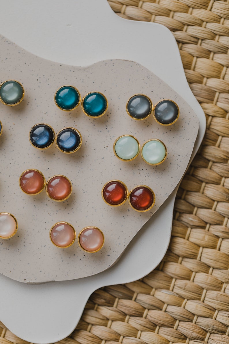May include: Assortment of colorful round gemstone earrings displayed on a white ceramic plate. The earrings are gold-toned and feature various colors, including blue, green, red, pink, and white.