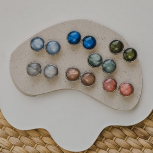 May include: Assortment of colorful round earrings with silver settings displayed on a white surface. The earrings are arranged in pairs and feature a variety of colors, including blue, green, brown, and pink.
