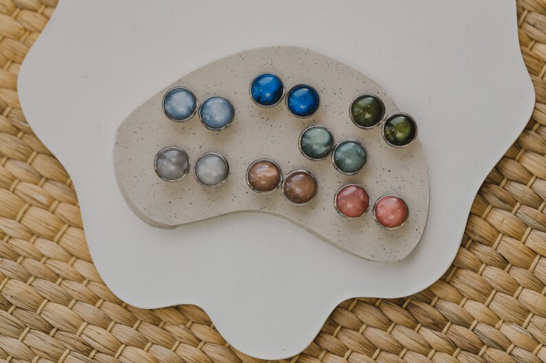 May include: Assortment of colorful round earrings with silver settings displayed on a white surface. The earrings are arranged in pairs and feature a variety of colors, including blue, green, brown, and pink.