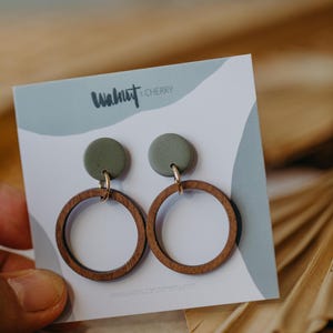 May include: A pair of green and brown wooden earrings with a round shape. The earrings are on a white card with the text "Walnut & Cherry" and a website address.